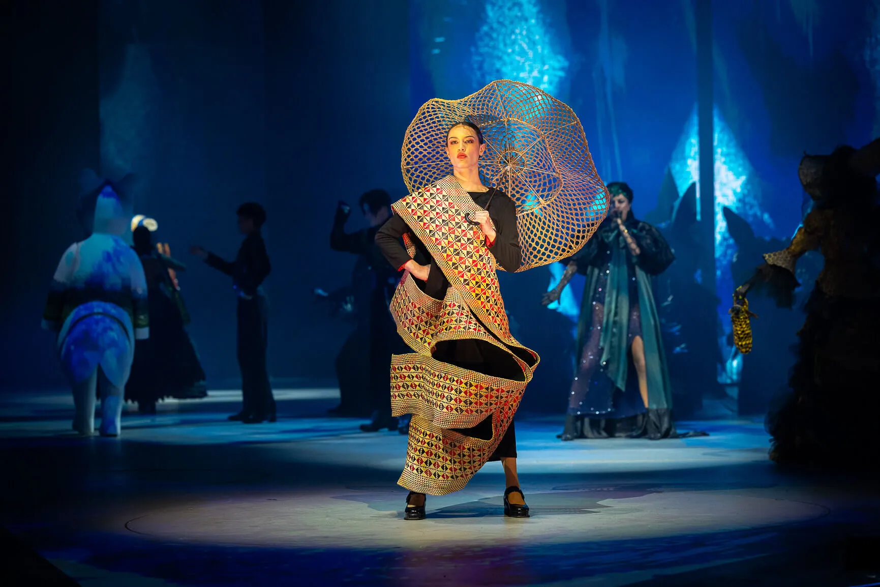 Model on runway wearing a large woven patterned wrap and circular woven headpiece with people and performers in the background under blue lighting.